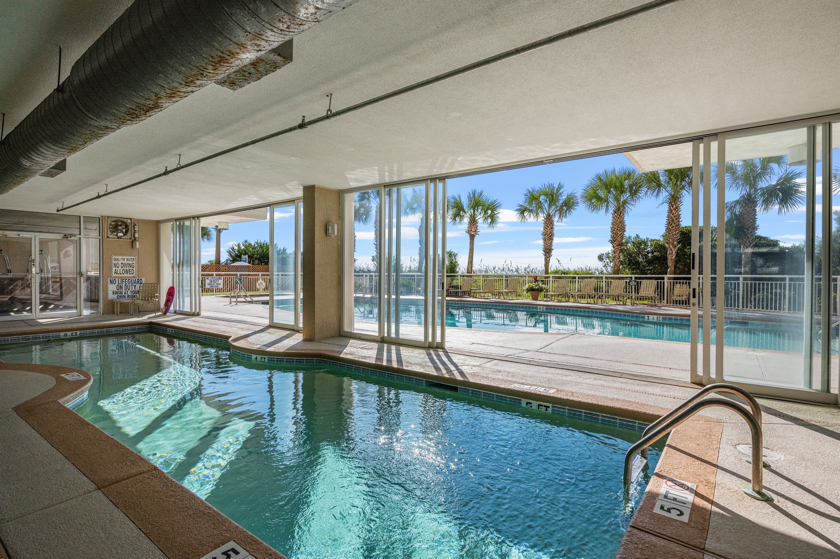 Una piscina interior con paredes de vidrio transparente, vistas a palmeras y un cartel de socorrista.