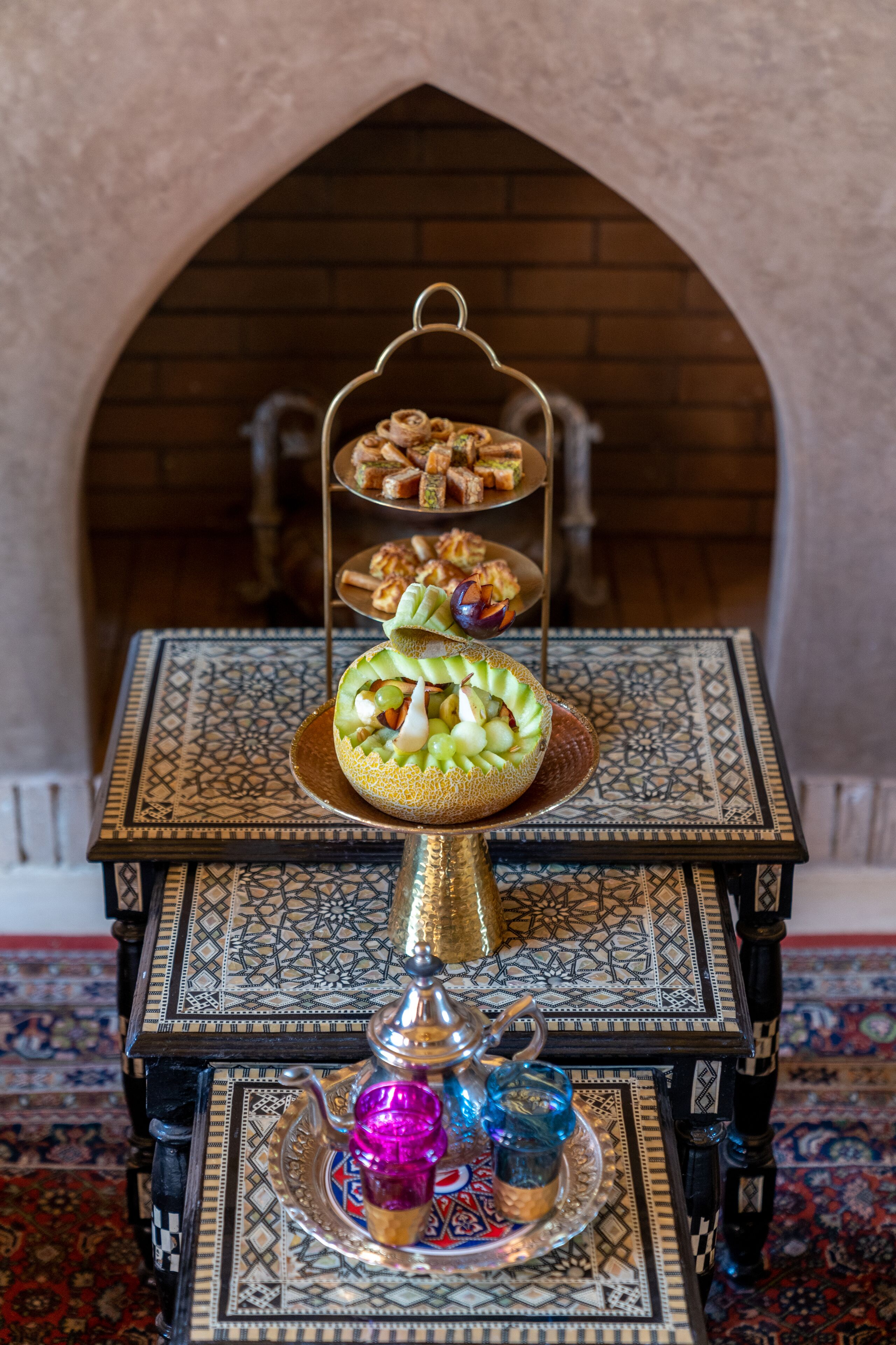 Un soporte de tres niveles para postres con diferentes pasteles y una pieza decorativa central sobre una mesa con elaborados diseños de azulejos.