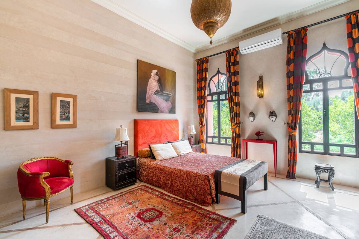 A bedroom with a large bed, a red armchair, a nightstand, and a red and orange patterned curtain.
