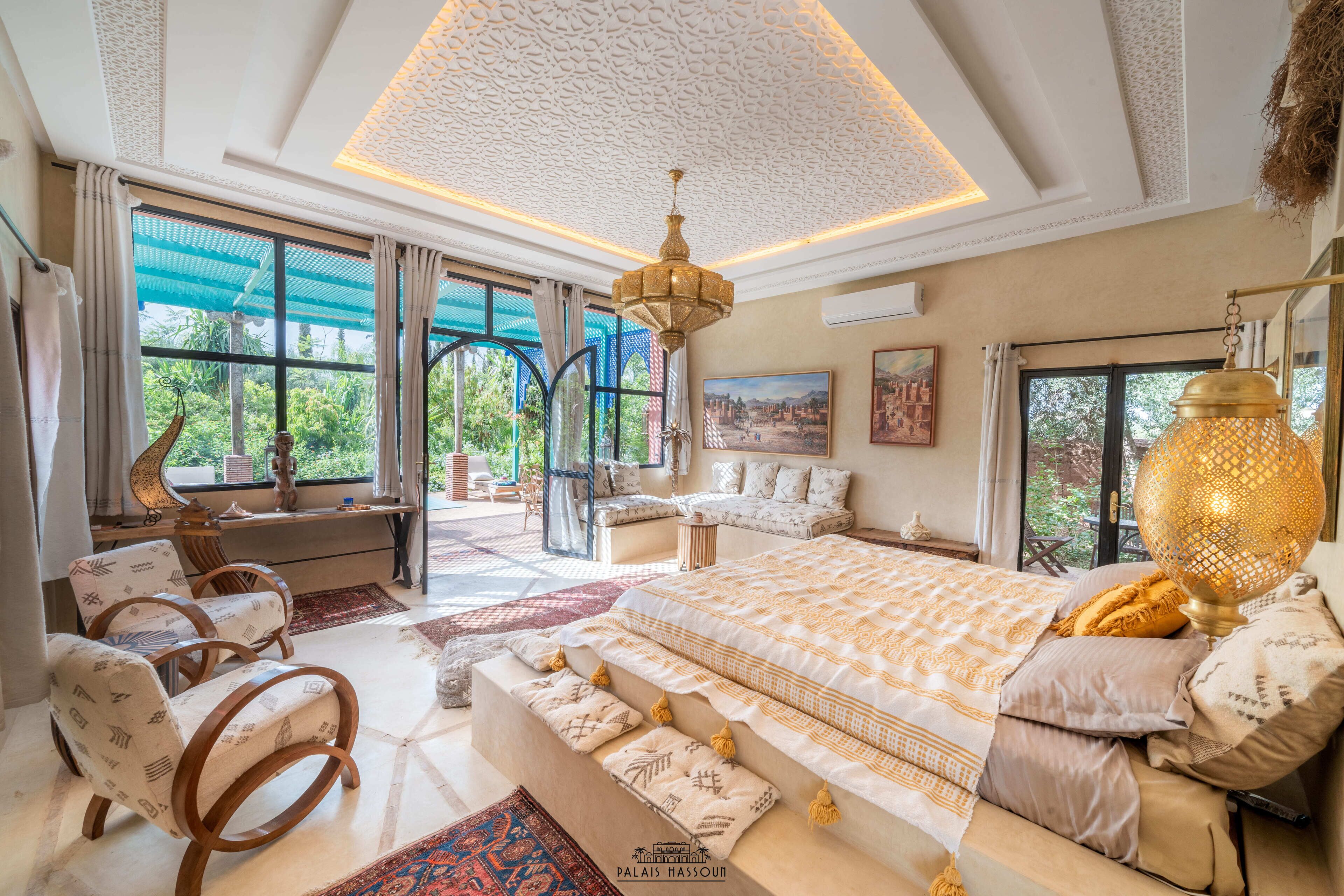 A spacious bedroom with a large bed, a sitting area with a sofa and chairs, and a view of greenery outside.