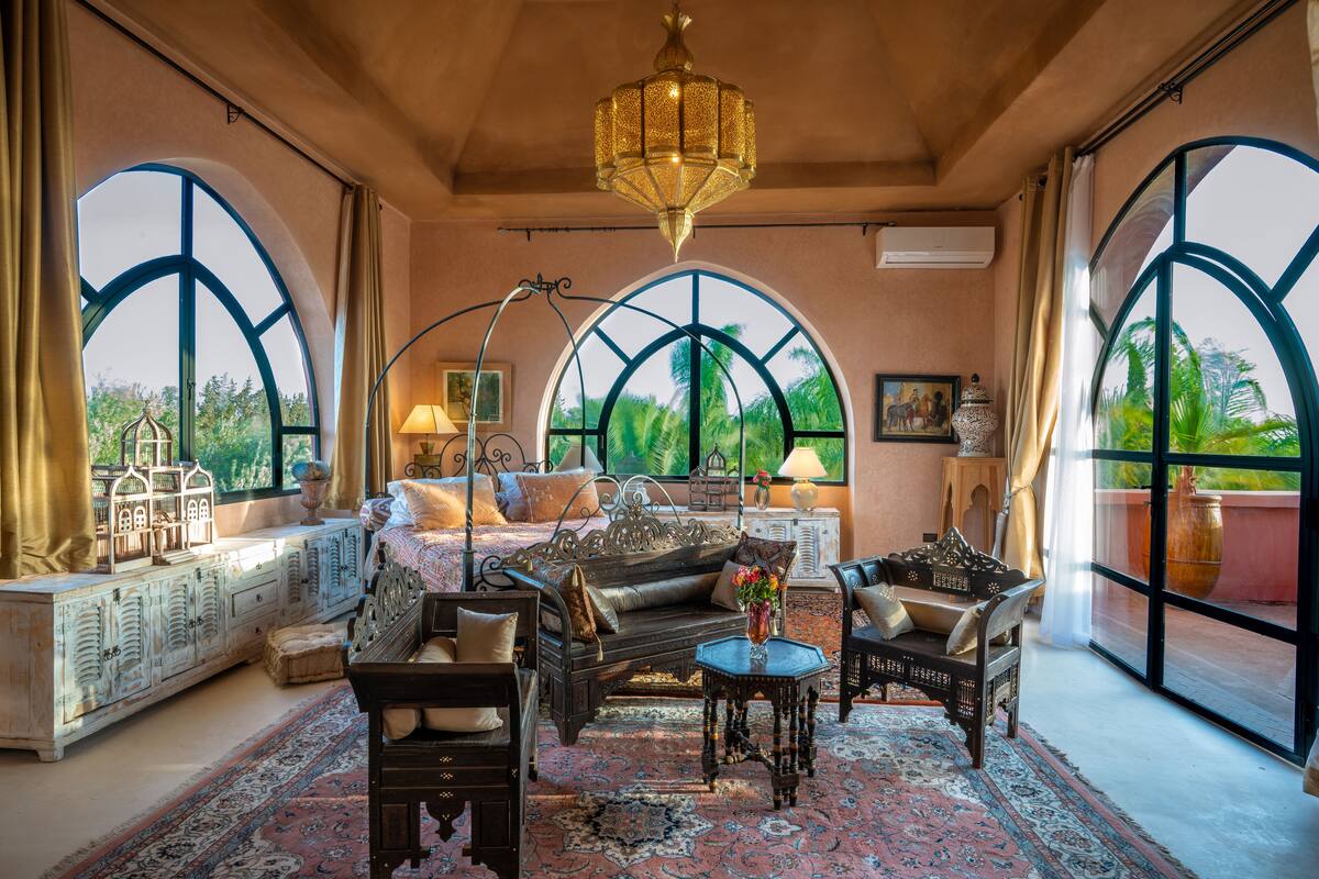 A spacious room with a large bed, a sofa, and two armchairs, a chandelier, and large windows with a view of greenery.