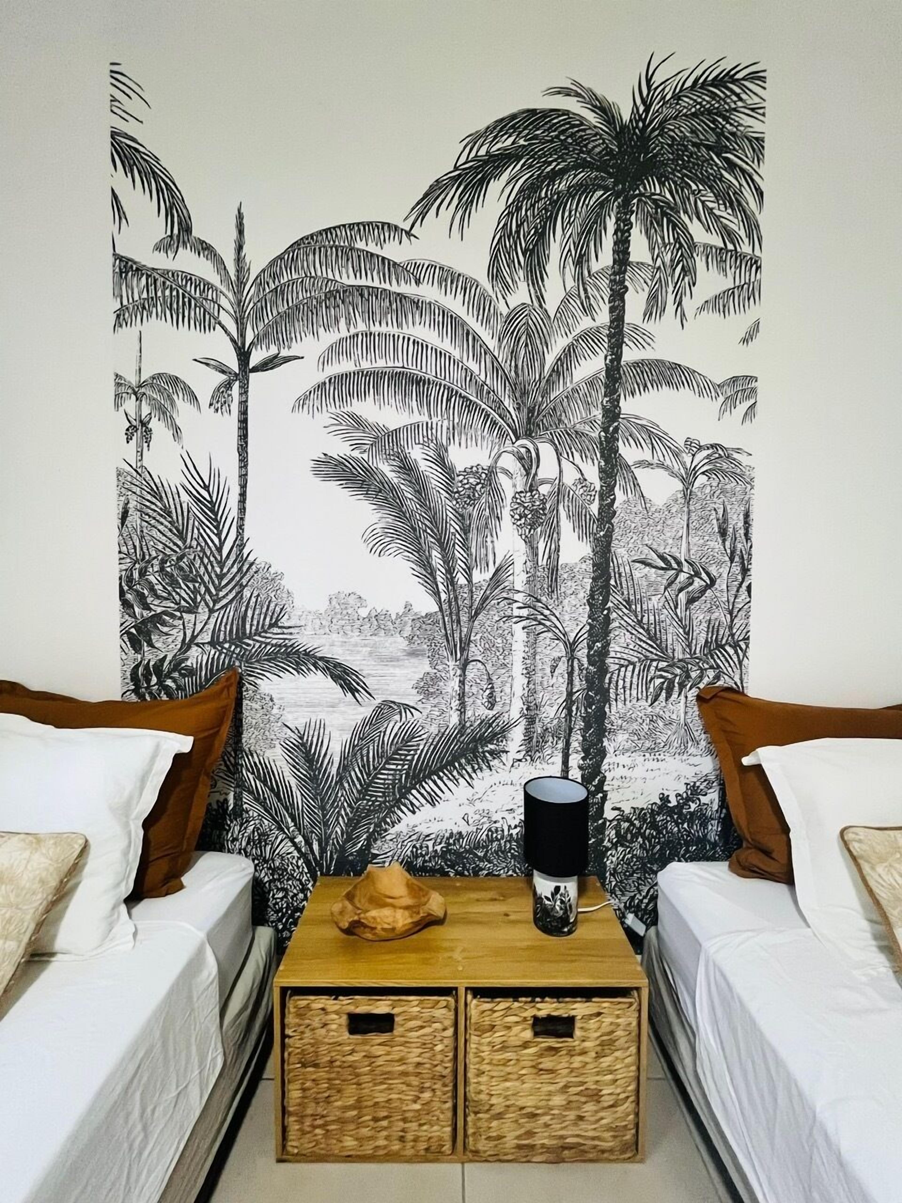 Una camera da letto con due letti singoli, un comodino in legno con una tazza nera e un arazzo con palme sulla parete.
