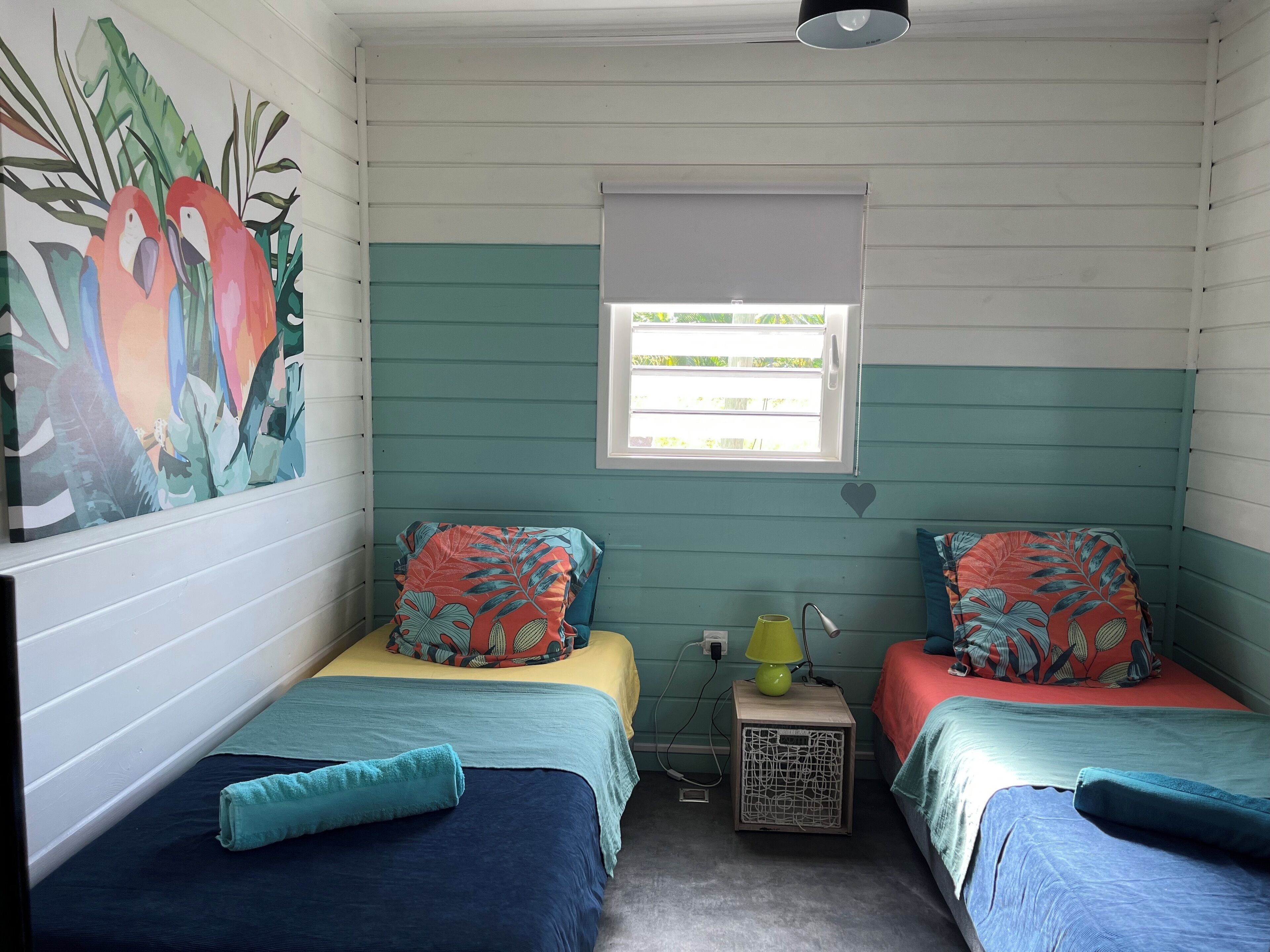 Una habitación con dos camas, cada una con una colcha floral de colores y almohadas a juego. Hay una mesita de noche con una lámpara, y una ventana que da a zonas verdes.