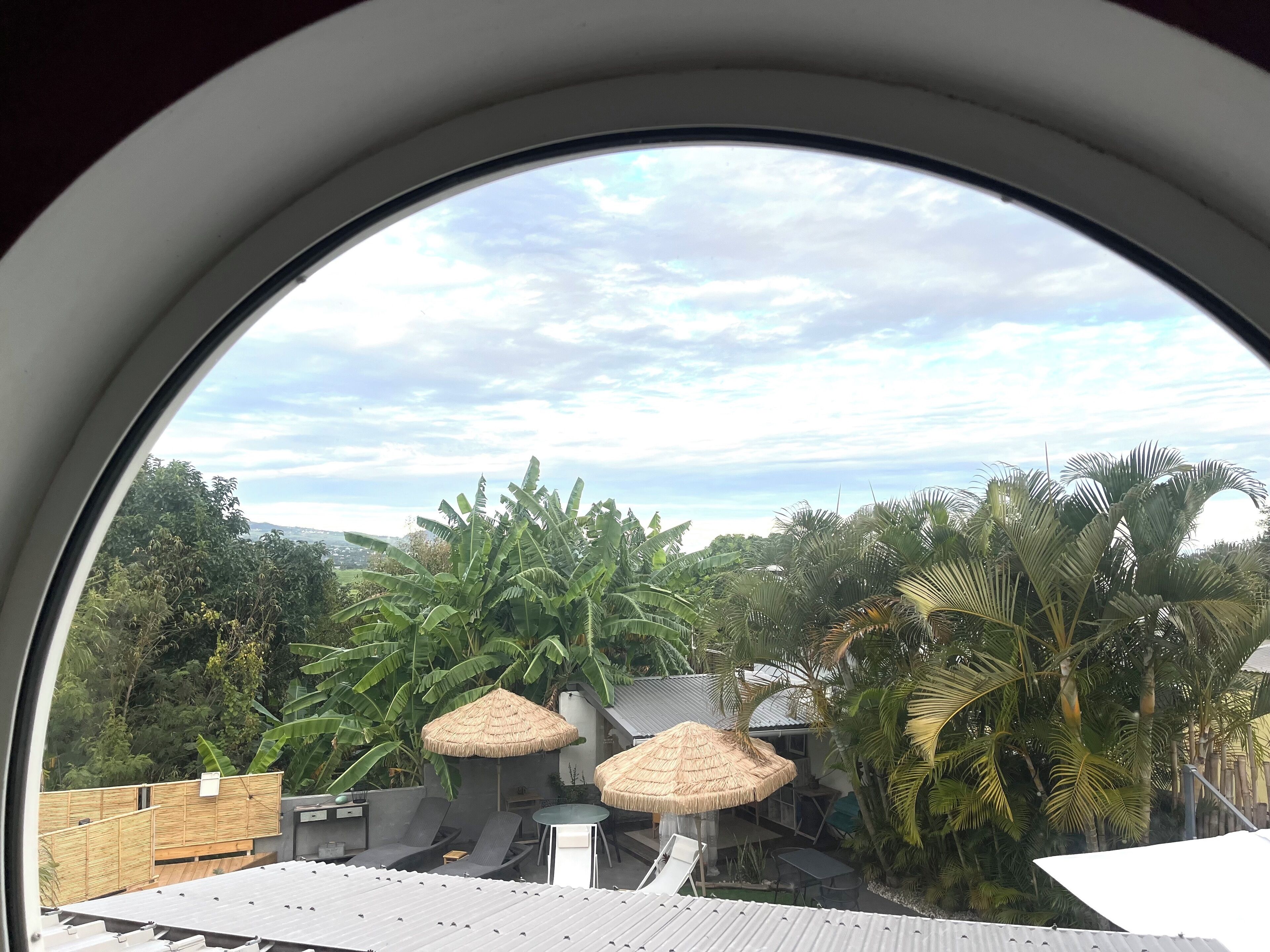 Blick durch ein Fenster auf eine Dachterrasse mit reetgedeckten Sonnenschirmen, Sitzgelegenheiten im Freien und üppiger tropischer Vegetation.