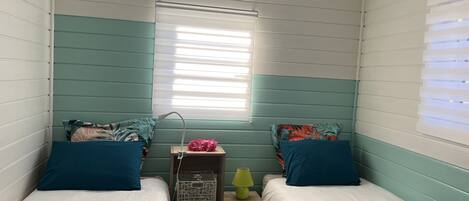 A room with two beds, each with teal bedding and matching pillows. There is a small table with a lamp and a decorative item.