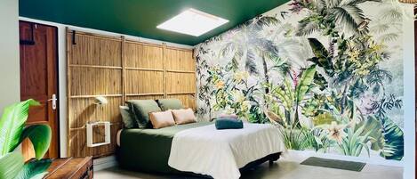 A bedroom with a bed, a bedside table, a lamp, and a large tropical mural on the wall.
