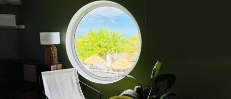 A room with a round window, a chair, a small stool, a lamp, and a potted plant.
