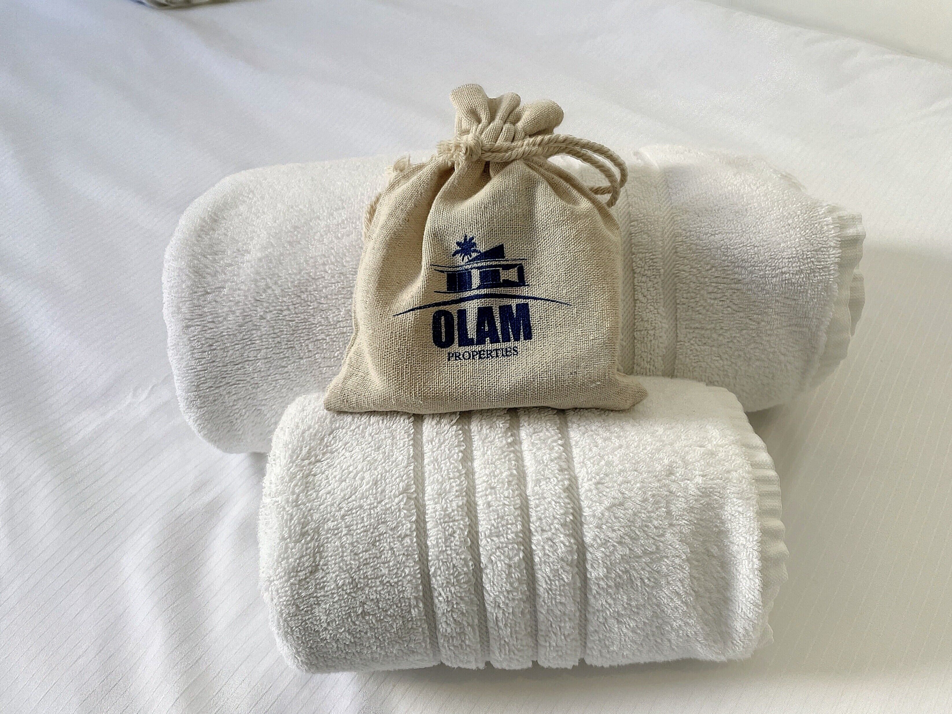 Une serviette blanche, soigneusement pliée, est posée sur un lit, accompagnée d’un sac en toile de jute muni d’un cordon de serrage, sur lequel est inscrit « OLAM PROPERDUES ».