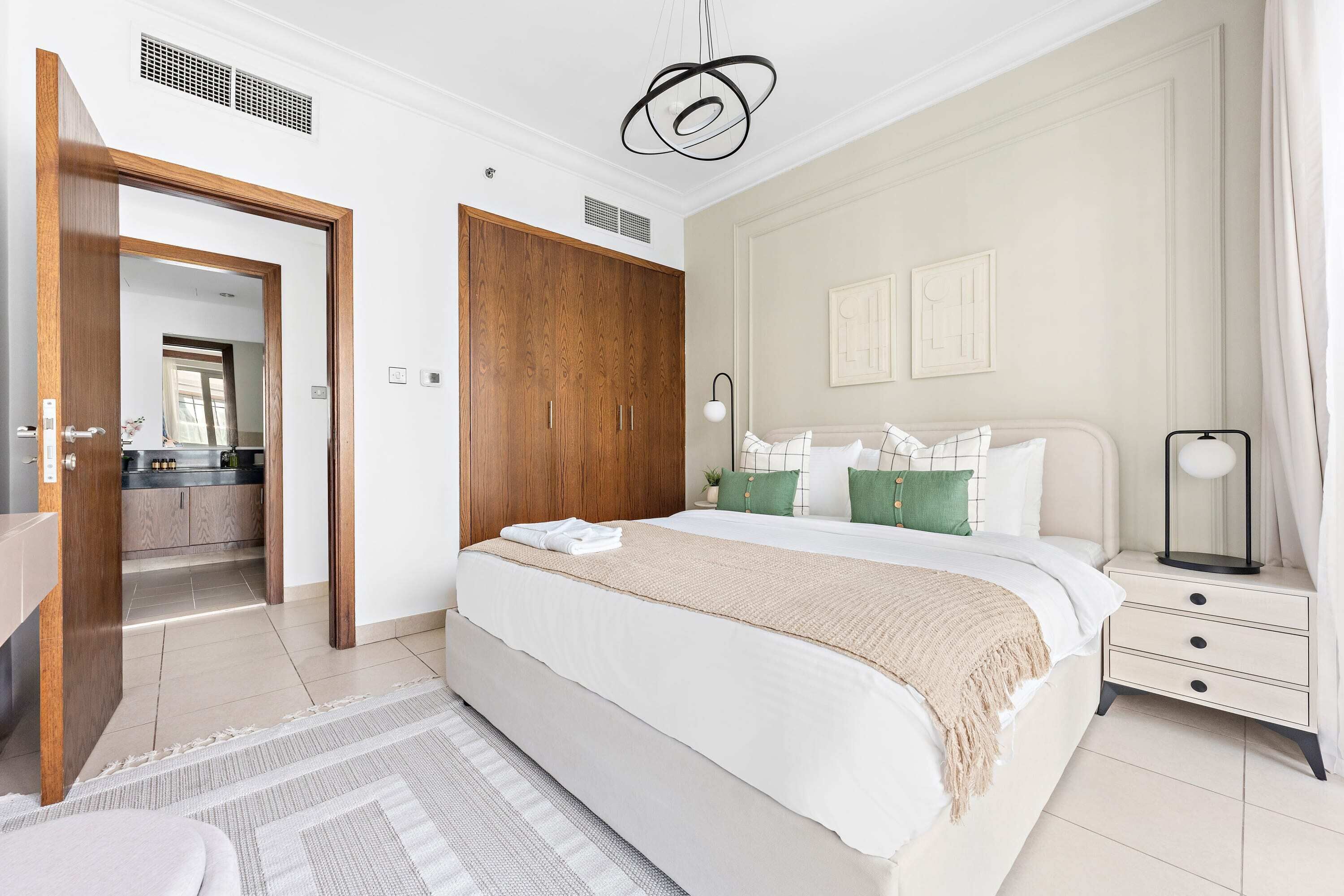 A bedroom with a large bed, a wooden wardrobe, a bedside table with a lamp, and a view into the kitchen.