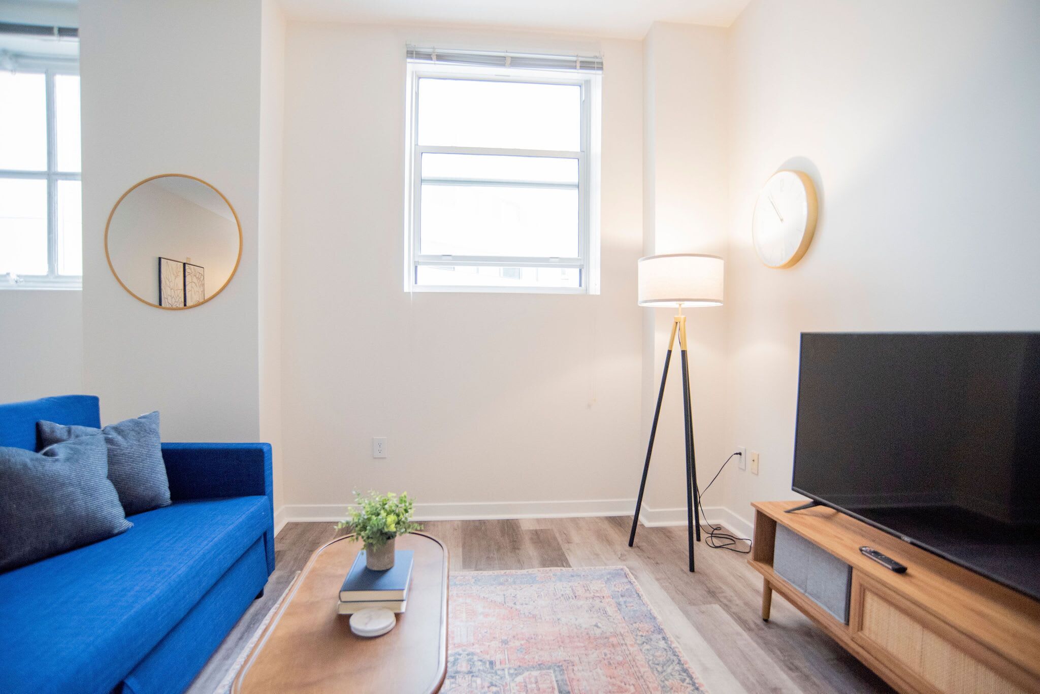 A living room with a blue sofa, a round mirror, a floor lamp, a television, and a window with blinds.