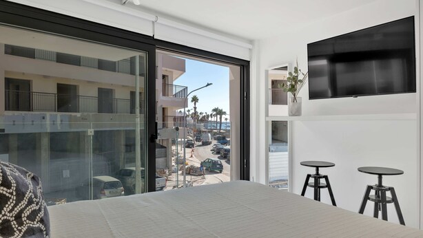A modern hotel room with a large bed, a TV mounted on the wall, and a balcony with a view of the street and other buildings. - Phaedrus Living: Alkminis Seaview Suites Pantai (Paphos)