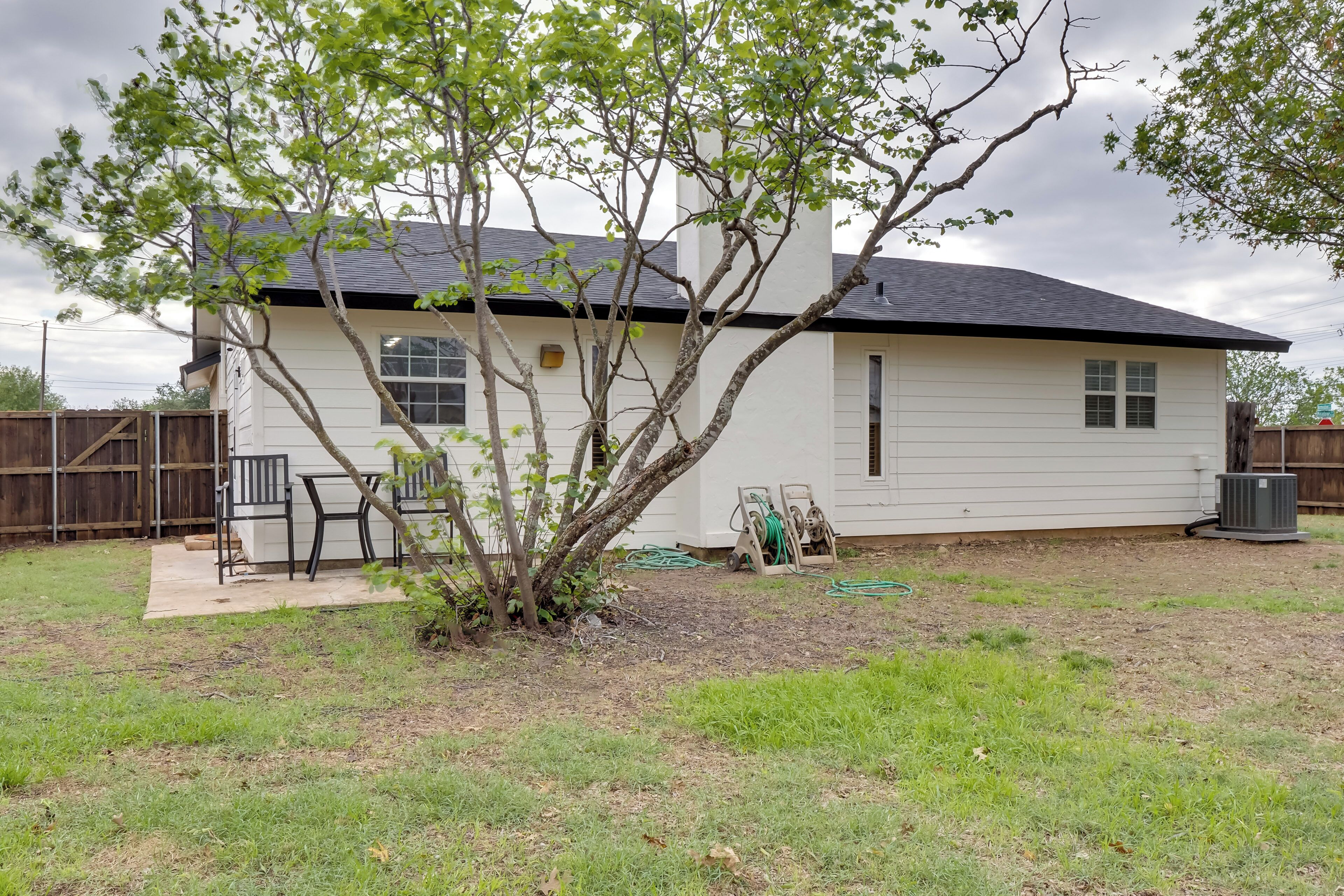 A single-story house with a white exterior and a dark roof. There is a small tree in the front yard, and a wooden fence is visible in the background.