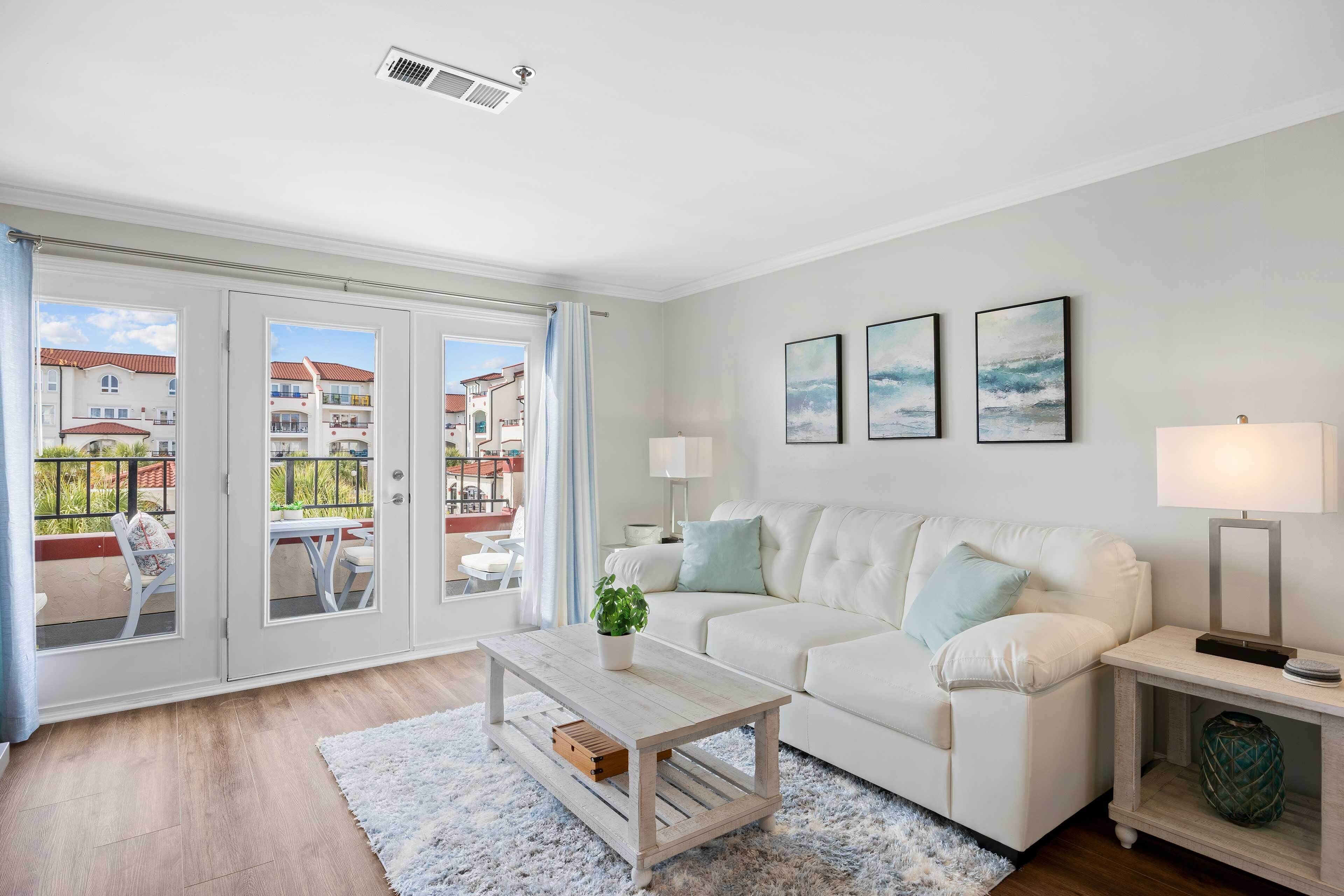 A modern living room with a white sofa, a coffee table, and a side table with a lamp. There are three framed pictures on the wall and a view of a balcony with outdoor furniture.
