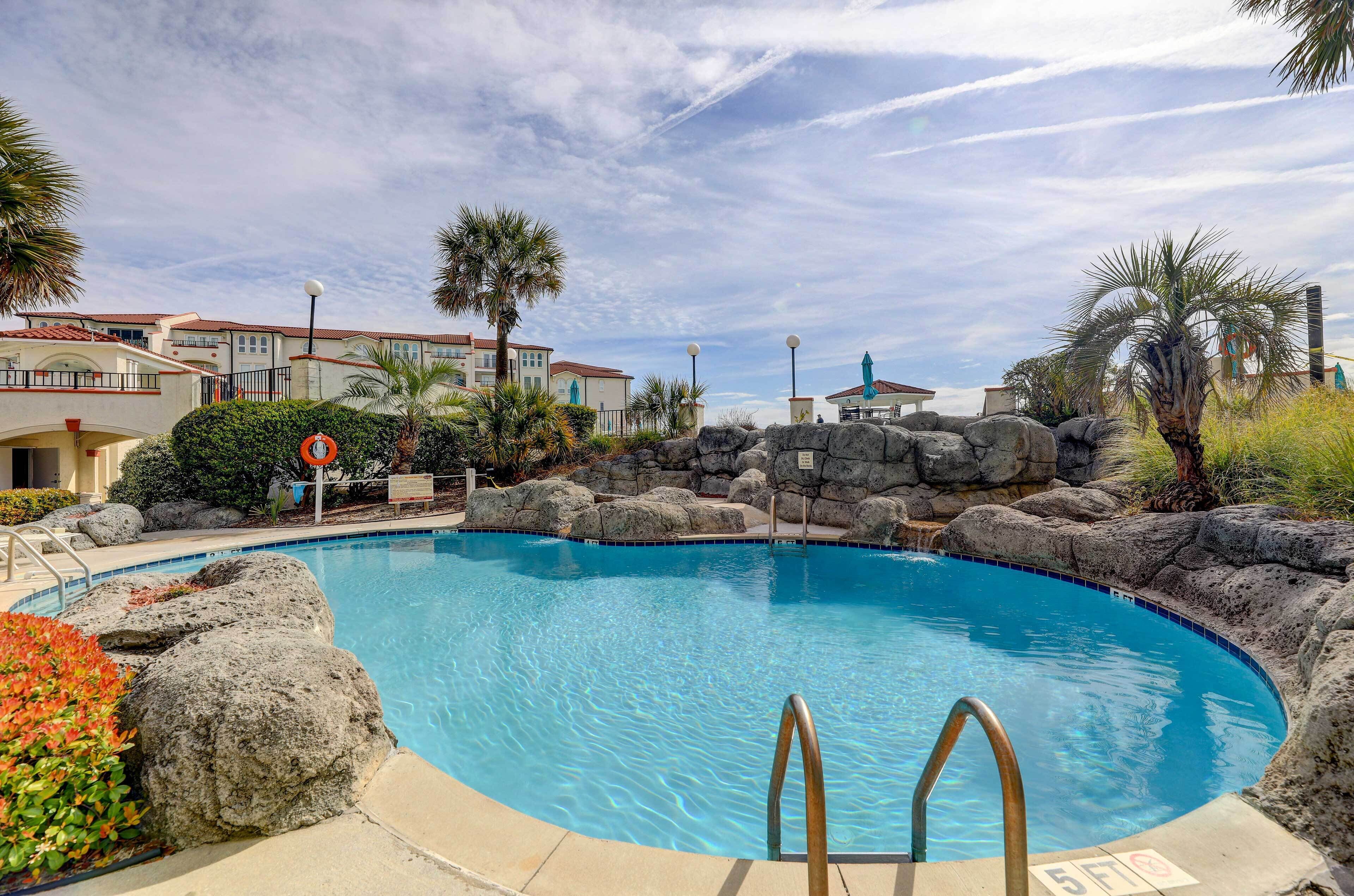 A swimming pool with a stone border, surrounded by palm trees and a building in the background.