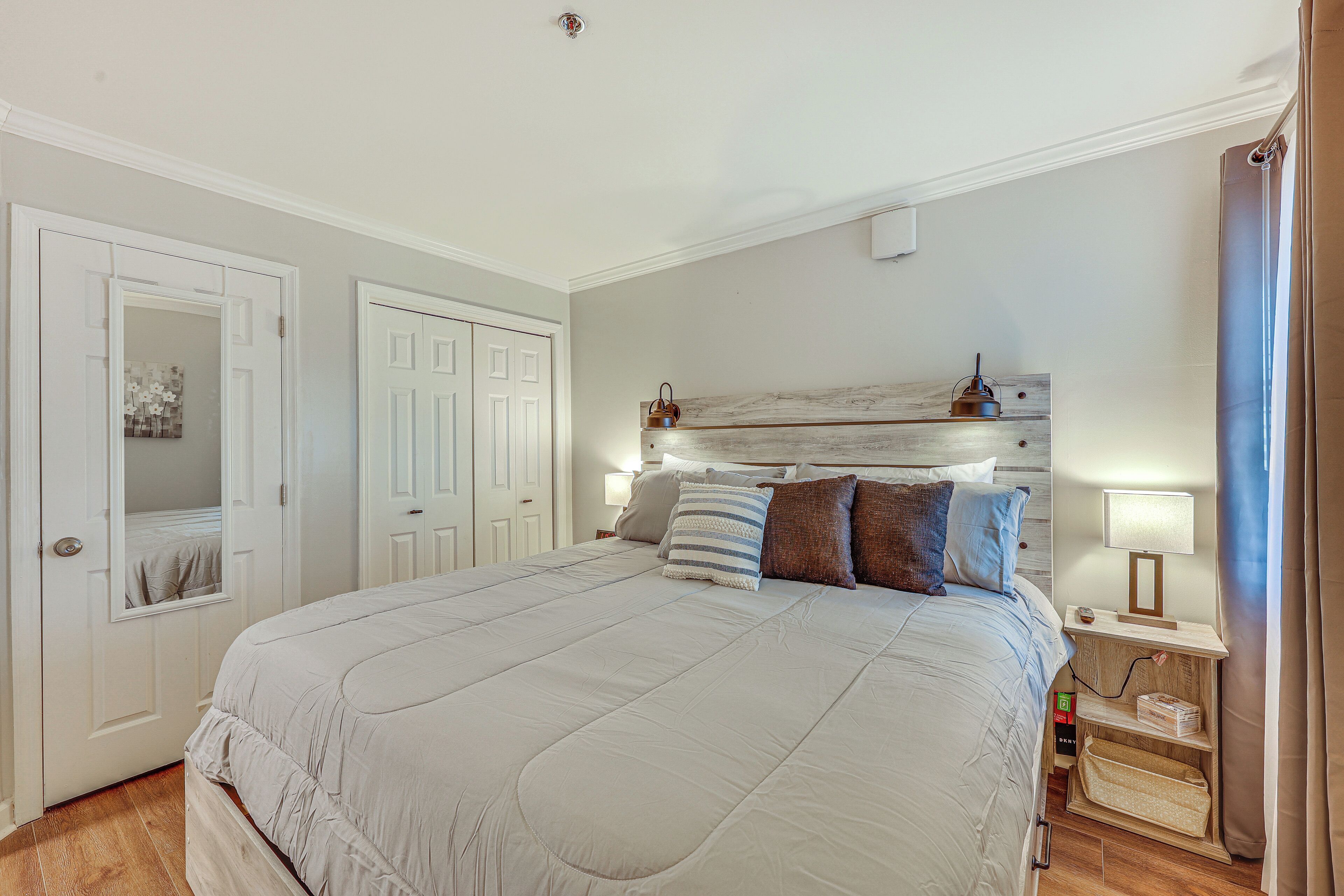 Un dormitorio ordenado con una cama grande, una mesita de noche con lámpara, y una superficie auxiliar con lámpara. Se observa una puerta con espejo y un estante de pared con objetos decorativos.