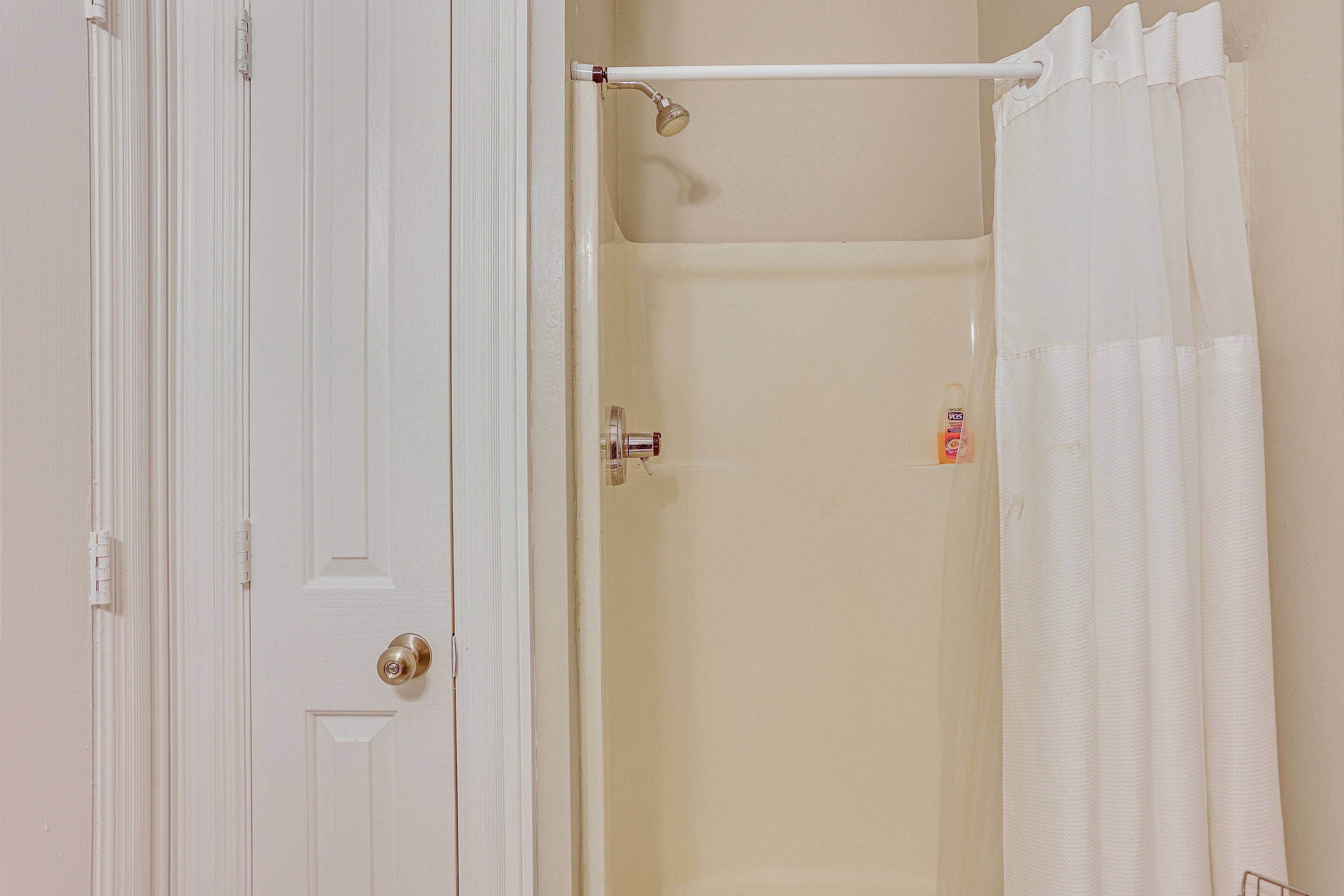 A bathroom with a white door, a shower curtain, and a visible bottle of shampoo.