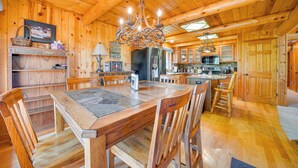 Ein Interieur einer Holzhütte mit einem Esstisch aus Steinplatten, Holzstühlen, einer Chandelieber und einer Küchenzeile mit Holdekor und Geräten.