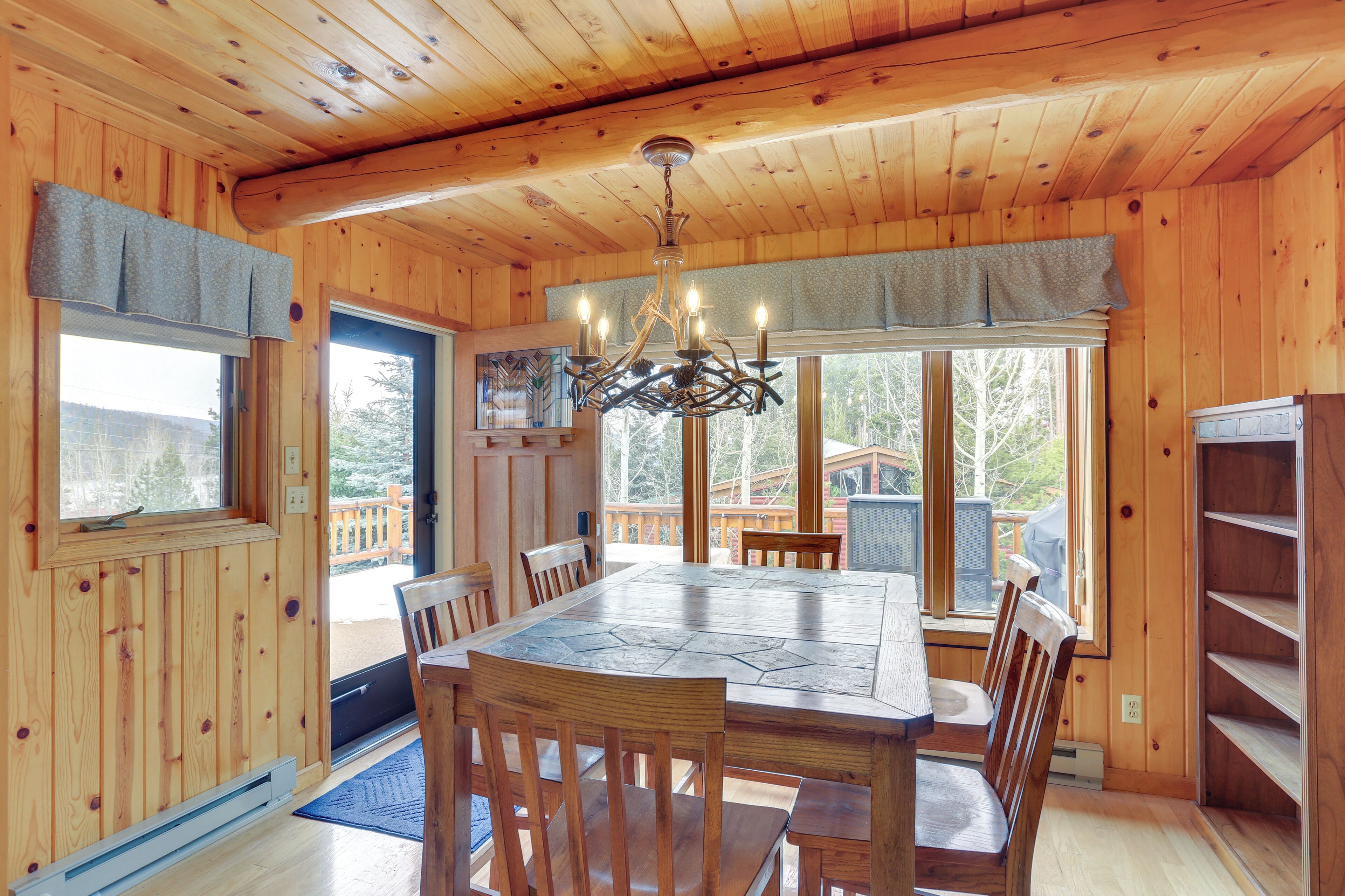 A wooden interior with a dining table, chairs, a chandelier, and large windows.
