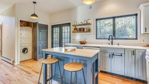 A modern kitchen with a central island, bar stools, a built-in washer and dryer, and large windows offering a view of the outdoors. - Secluded Kerhonkson Retreat With Deck + Views! (Kerhonkson)