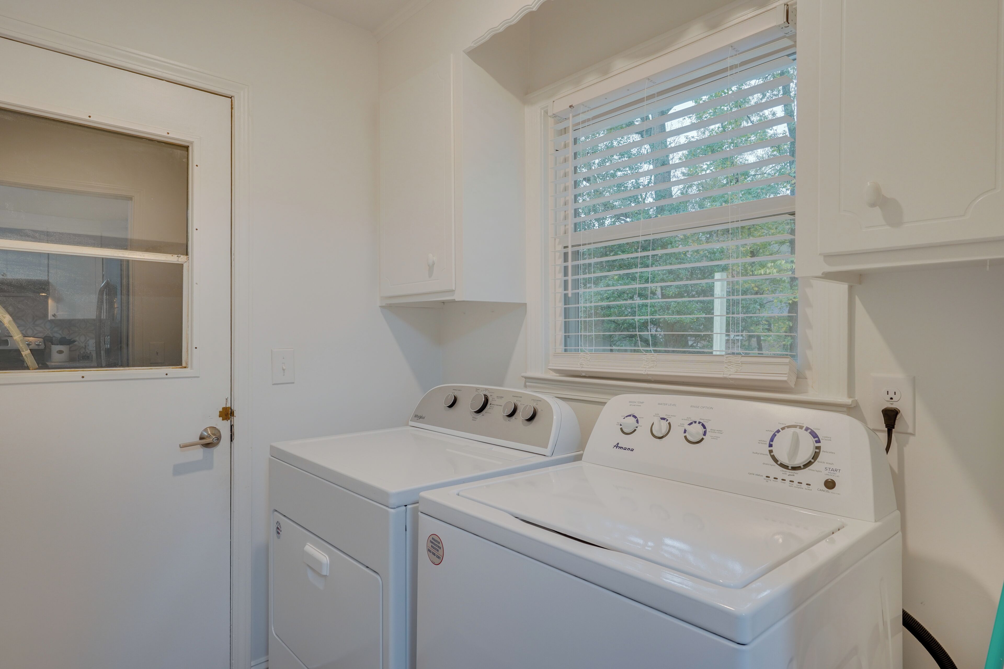 A laundry room with two white washers and dryers, a window with blinds, and a door with a glass panel.