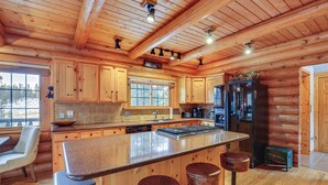 A log cabin kitchen with wooden cabinets, a central island with stools, a refrigerator, and a window overlooking a snowy landscape. - 4 Mi to Rocky Mtn Nat'l Park: Cabin Near Town! (Grand Lake)