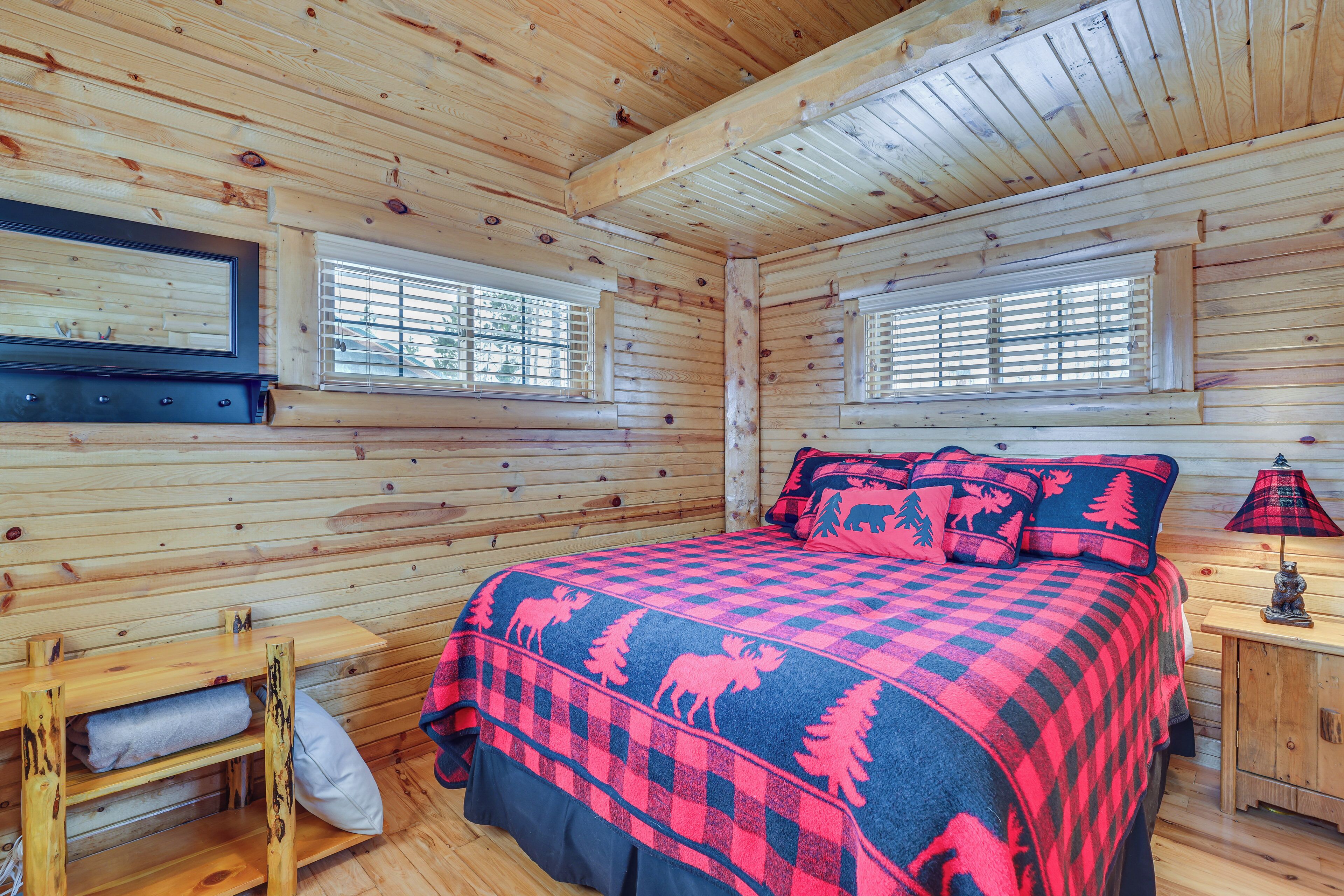 A log cabin bedroom with a checkered bedspread, wooden walls, a bench, and a window with blinds.