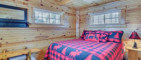 A log cabin bedroom with a checkered bedspread, wooden walls, a bench, and a window with blinds.