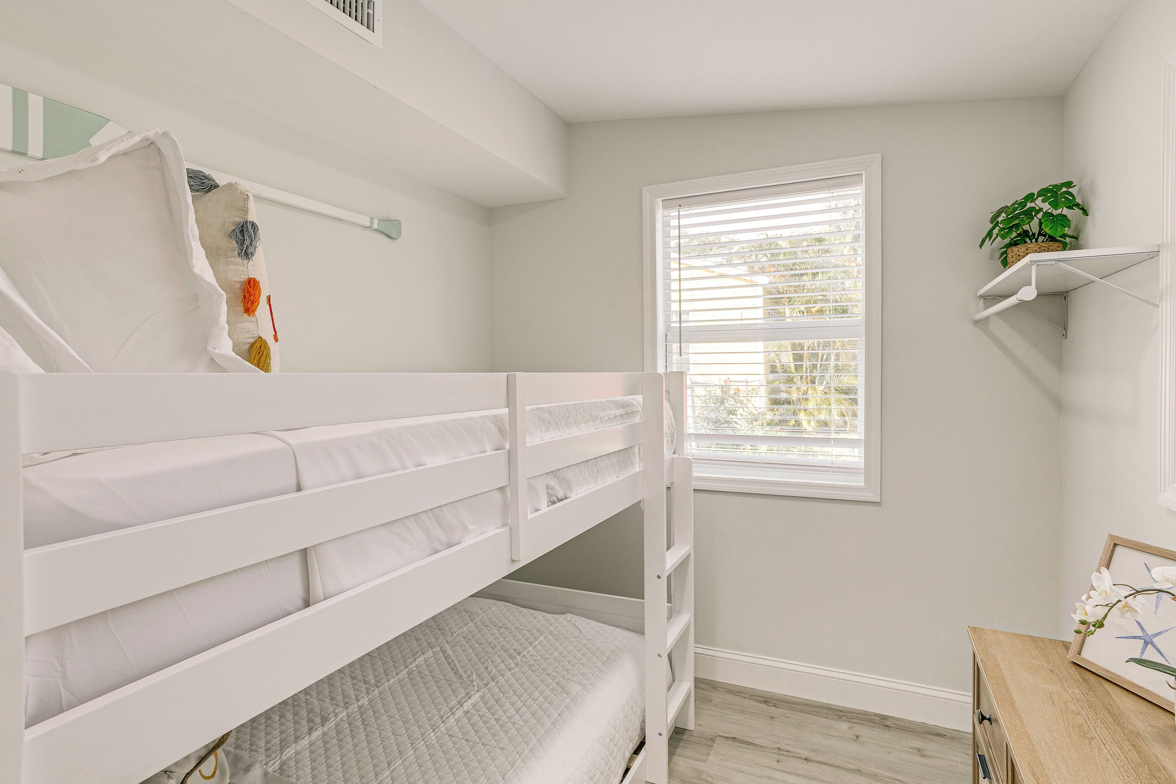 A child's bunk bed in a room with a window, a small wooden dresser, and a shelf with a plant.