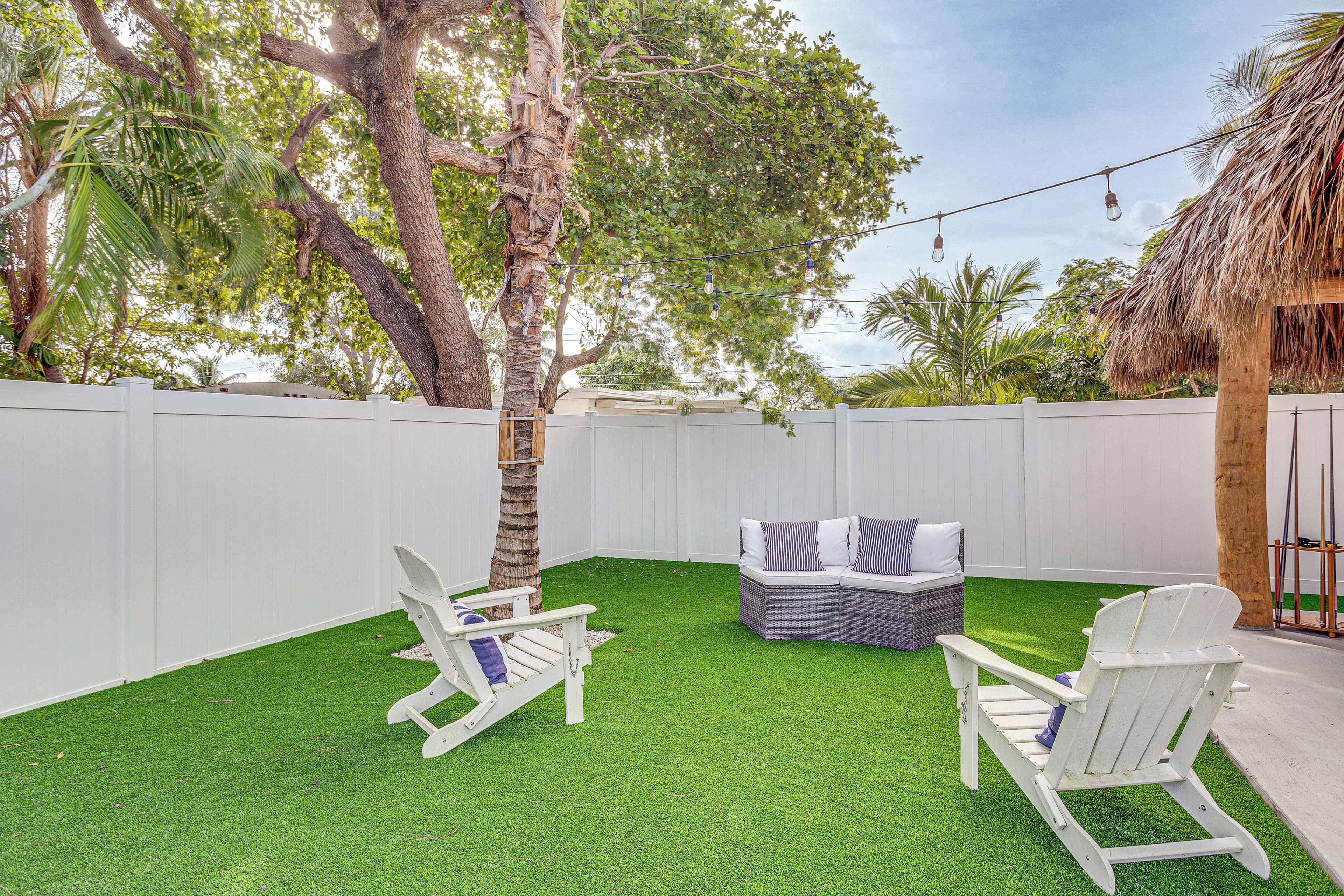 A backyard with artificial grass, two Adirondack chairs, a wicker sofa, and a thatched-roof structure.