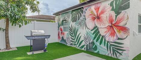A backyard with artificial grass, a barbecue grill, and a wall mural of tropical flowers.