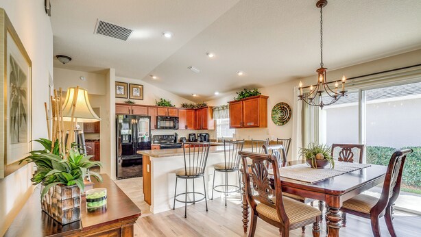 A modern kitchen with wooden cabinets, a central island with bar stools, and a dining area with a wooden table and chairs. - Screened Lanai & Grill: Home in The Villages (Wildwood)