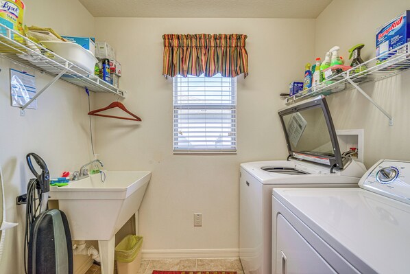 A laundry room with a washing machine, dryer, shelves with cleaning supplies, a sink, and a window with blinds. - Screened Lanai & Grill: Home in The Villages (Wildwood)