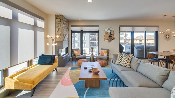 A modern living room with a sofa, a coffee table, and a bench. There are two chairs, a dining table, and a large window with a view of buildings. - Winter Park Condo w/ Hot Tub, Balcony & Shuttle! (Winter Park)