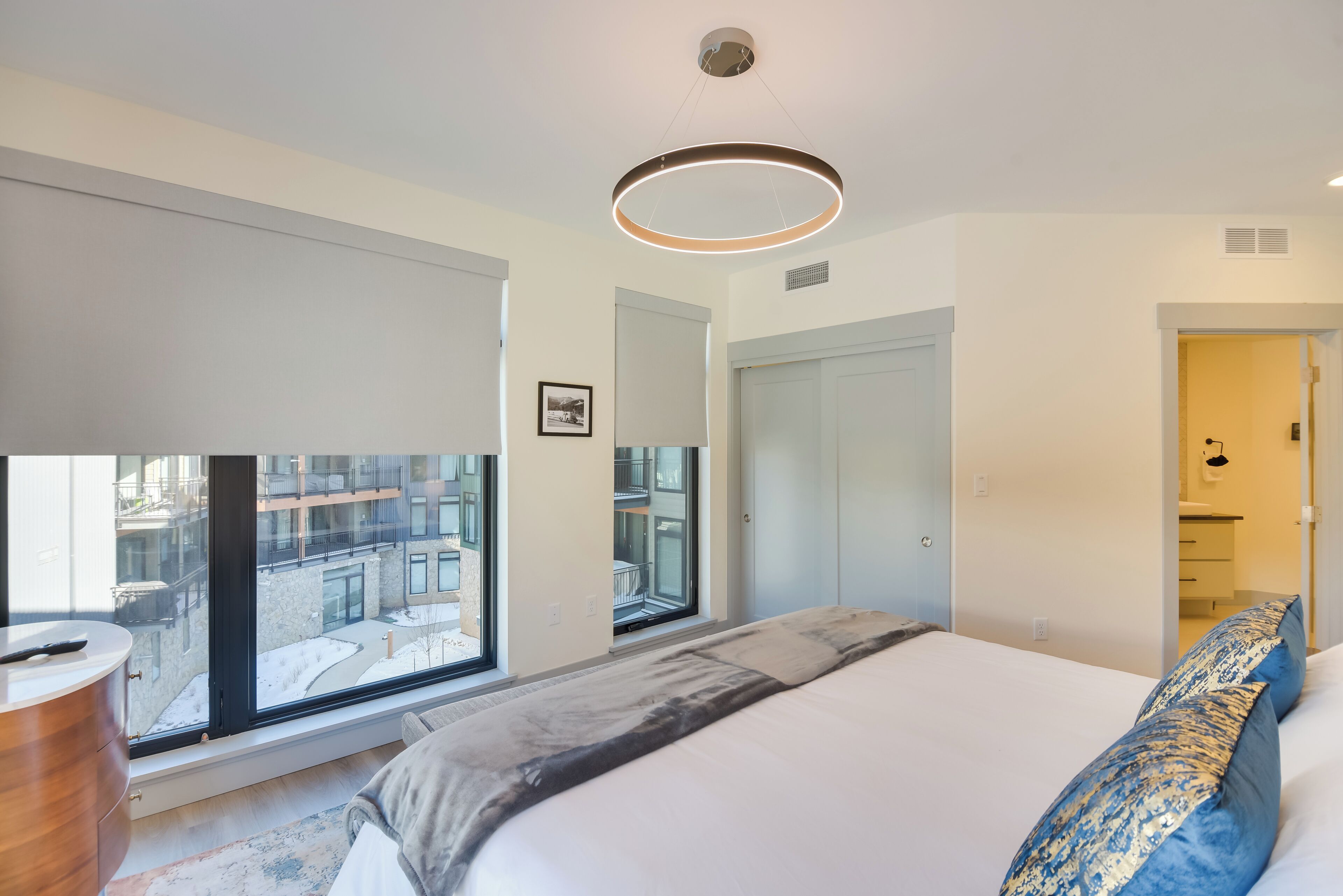 Une chambre moderne avec un grand lit, un luminaire circulaire au plafond et une vue sur le paysage urbain grâce à de grandes fenêtres.