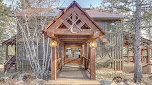 A wooden lodge with a covered walkway leading to the entrance, surrounded by trees and a rustic setting. - Ride to Sturgis! Lead Home w/ Pool Access (Lead)