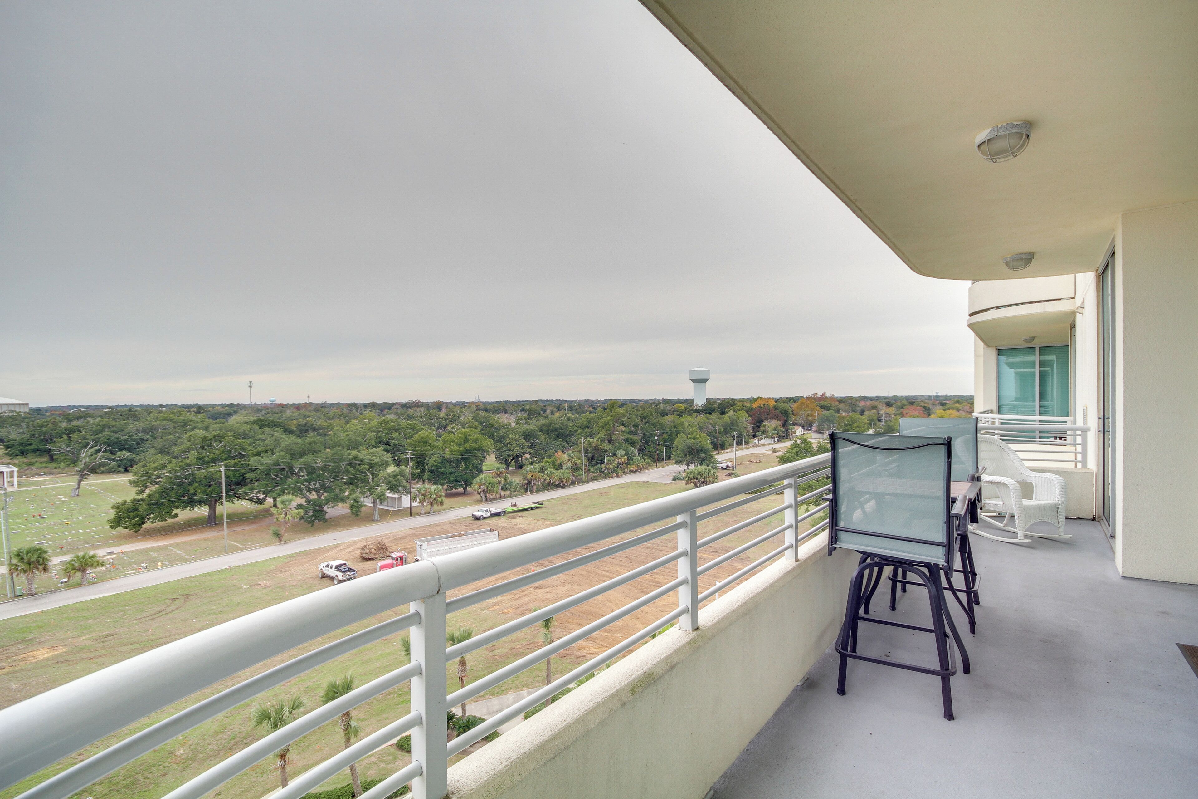 A balcony with a glass table and chairs, overlooking a green landscape with trees and a distant water tower.