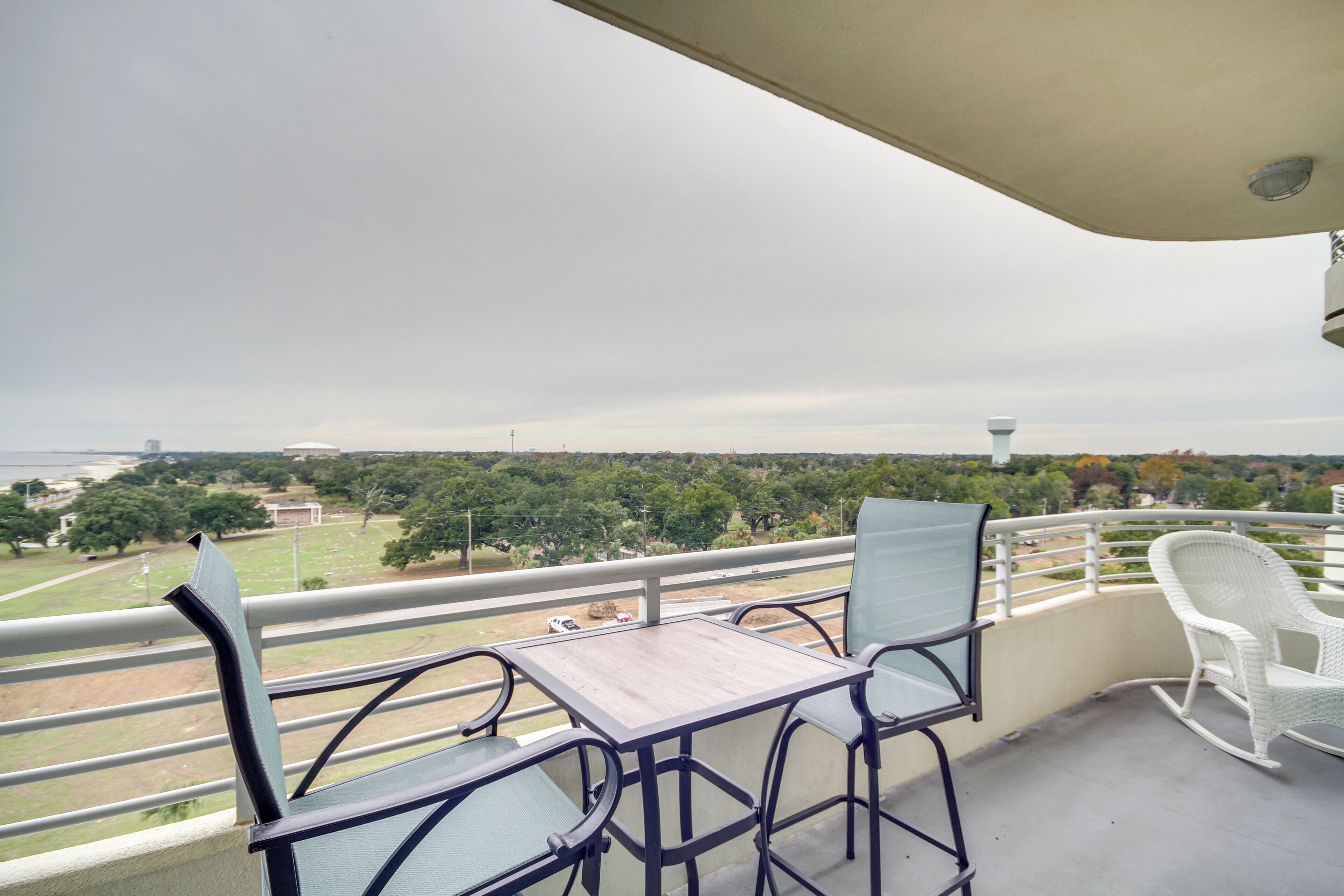 Un balcon avec une table et deux chaises, donnant sur un paysage verdoyant, avec des arbres et une tour qui se profile à l’horizon.