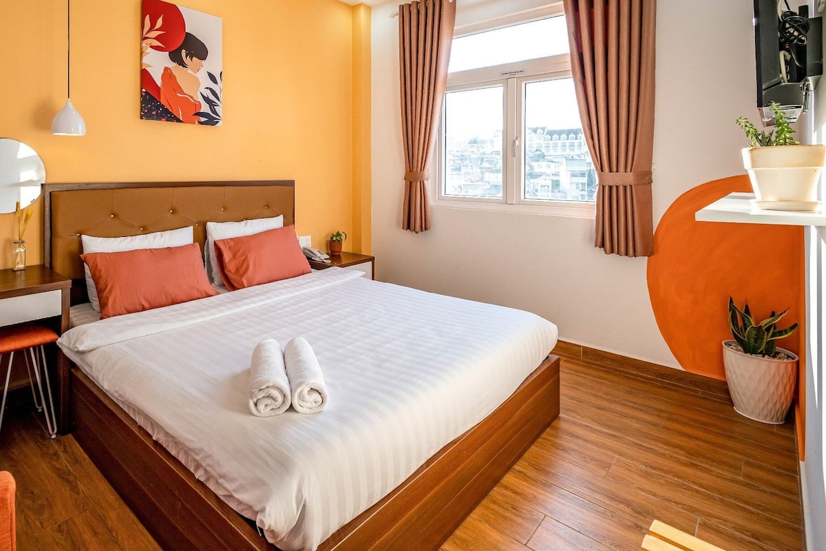 A hotel room with a bed, a desk, a chair, a mirror, a window with curtains, and a potted plant.