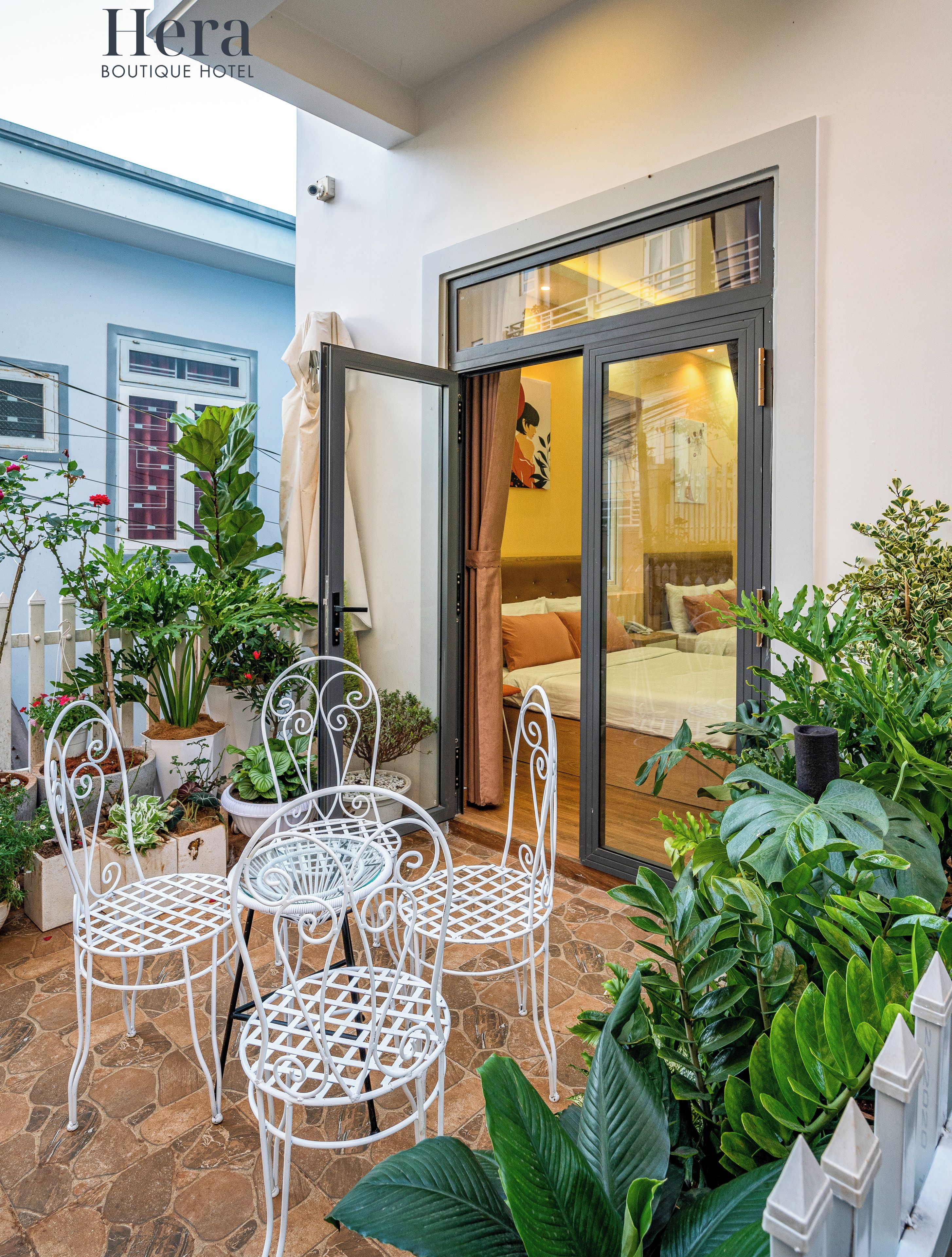A hotel patio with white metal chairs, potted plants, and a glass door leading to a room.