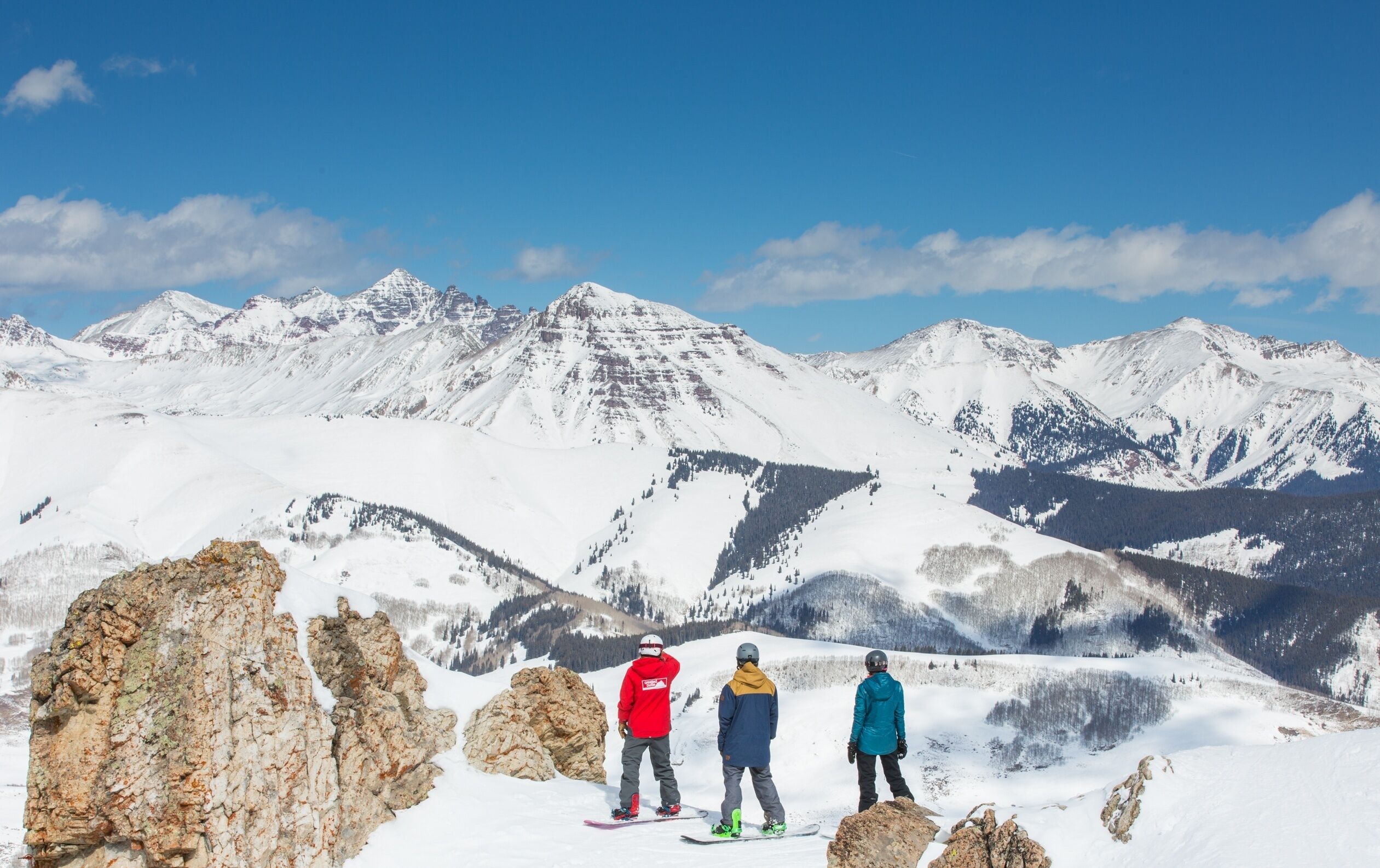 Paisaje nevado de montaña con tres esquiadores de pie sobre un saliente rocoso.