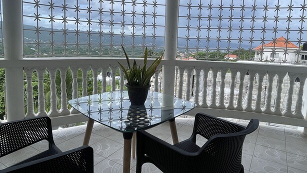 Mesa com tampo de vidro, duas cadeiras, um vaso de planta e vista para a paisagem costeira através de uma cerca de metal decorativa.