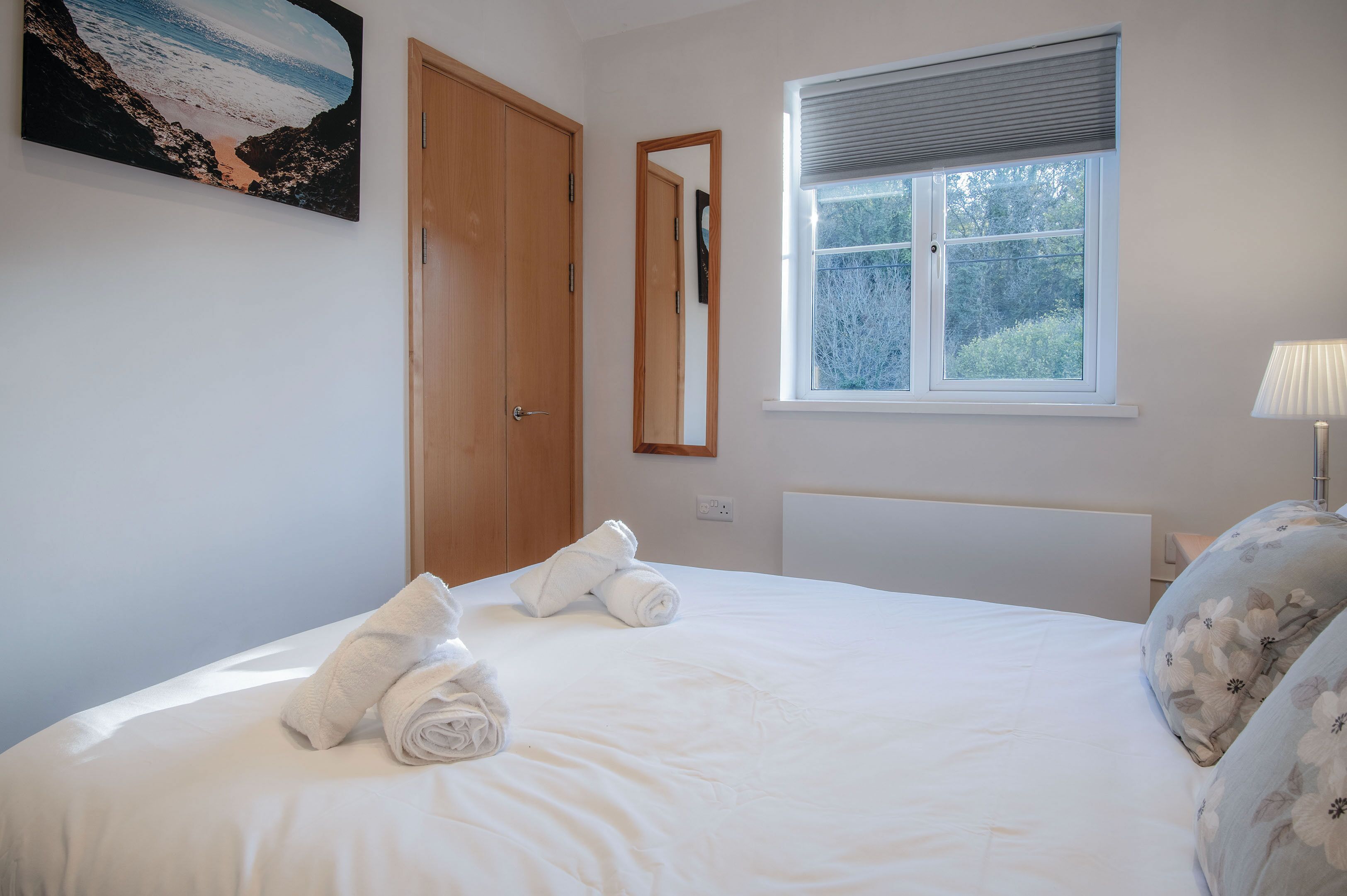 A neatly made bed with rolled towels, a wooden door, a window with blinds, and a wall-mounted mirror.