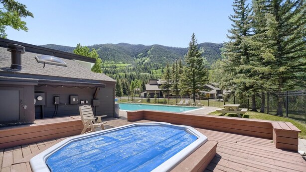 A hot tub on a wooden deck with a pool in the background, surrounded by a fence and trees.