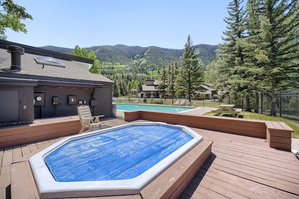 Un jacuzzi en una terraza de madera, con una piscina al fondo, rodeado por una valla y árboles.