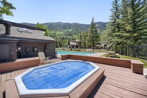 Un jacuzzi en una terraza de madera, con una piscina al fondo, rodeado por una valla y árboles.