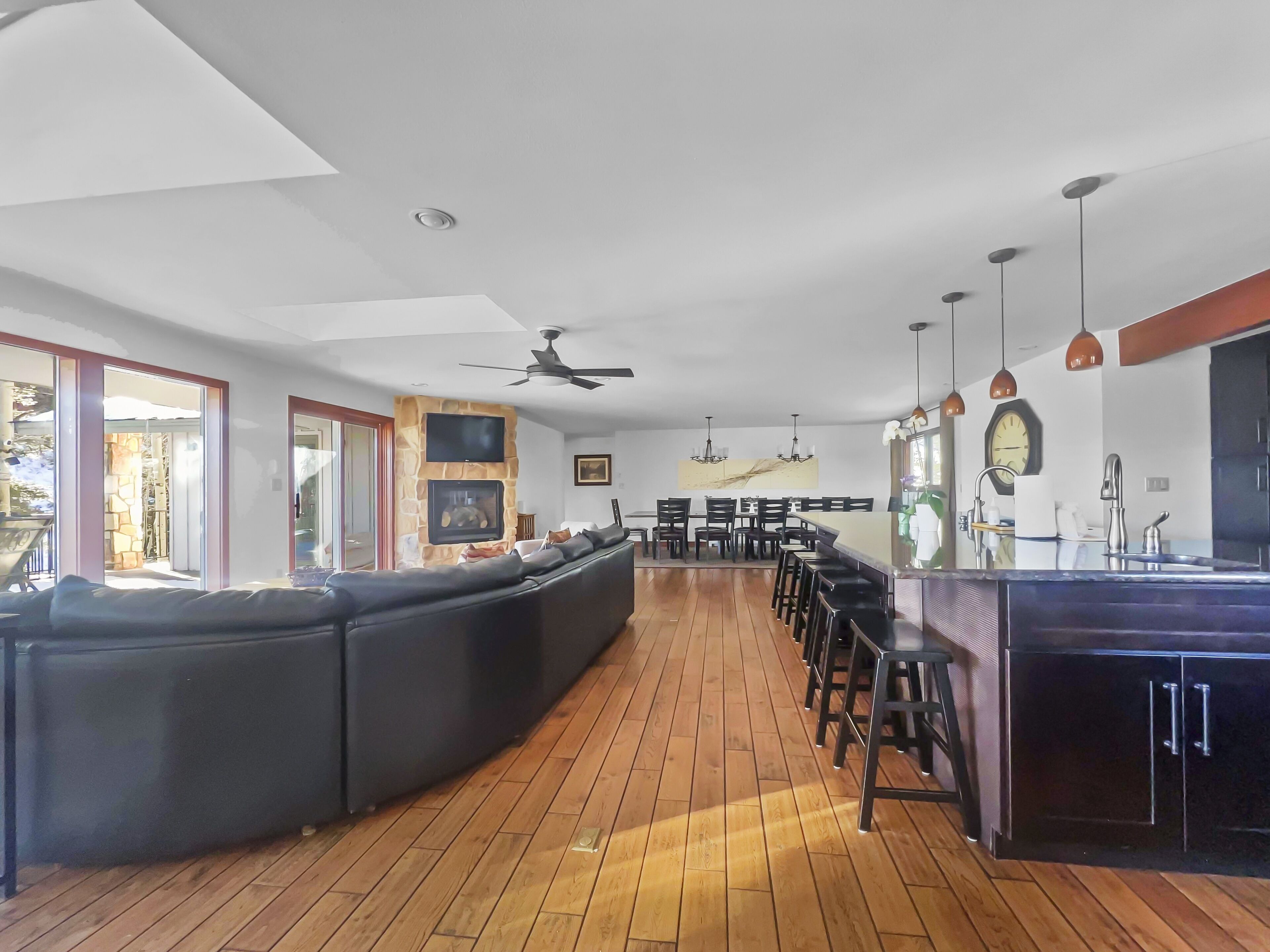 A modern kitchen with a built-in island, a dining area with a long table, and a living area with a large black sectional sofa.