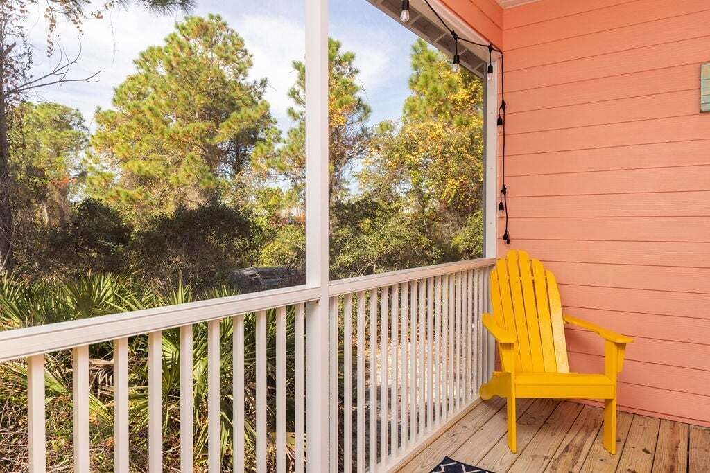 A wooden deck with a white railing, a yellow Adirondack chair, and a view of trees and foliage.
