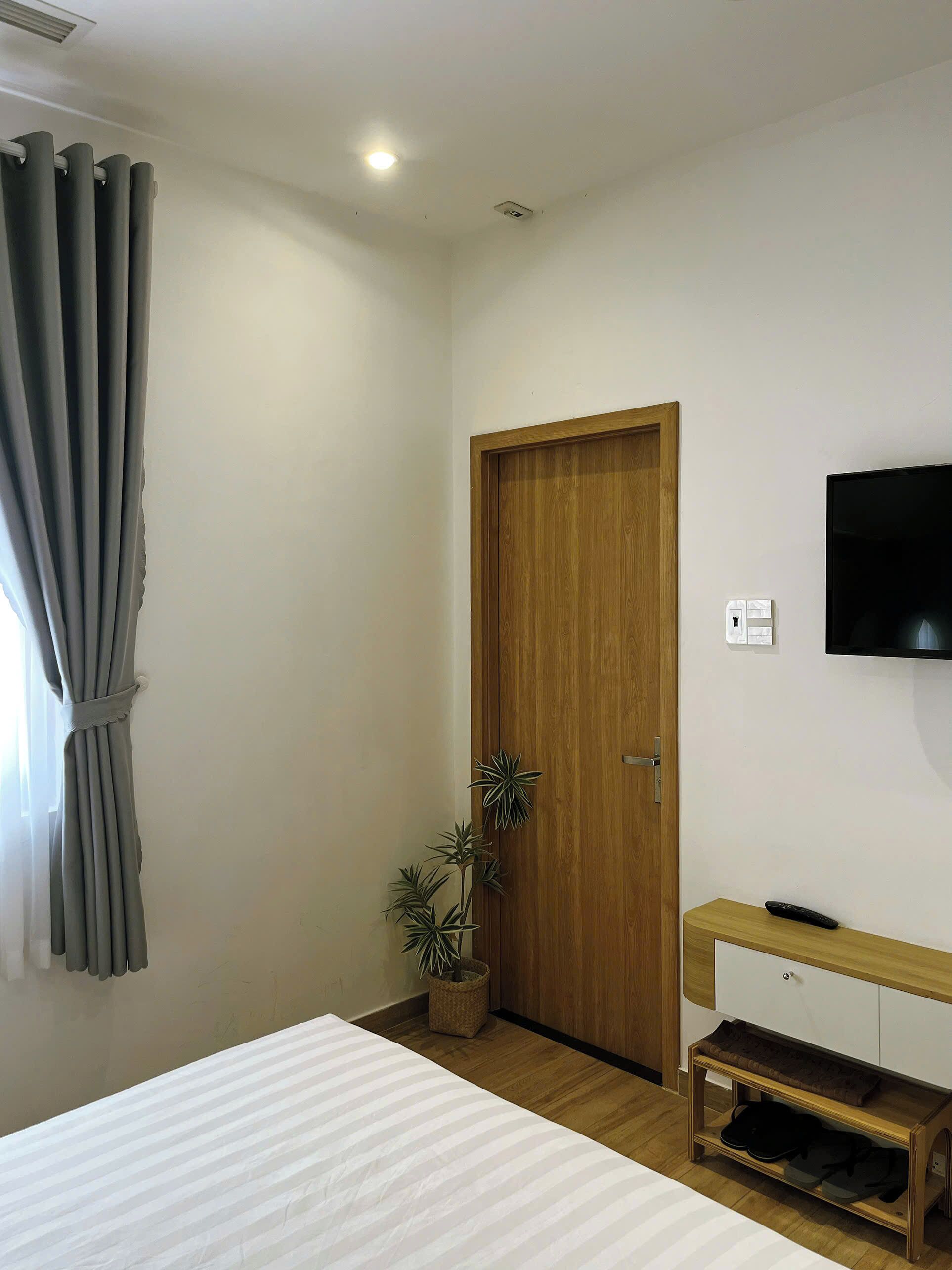 Un dormitorio con una cama, una puerta de madera, un televisor montado en la pared, una planta pequeña y una mesita de noche con un control remoto.