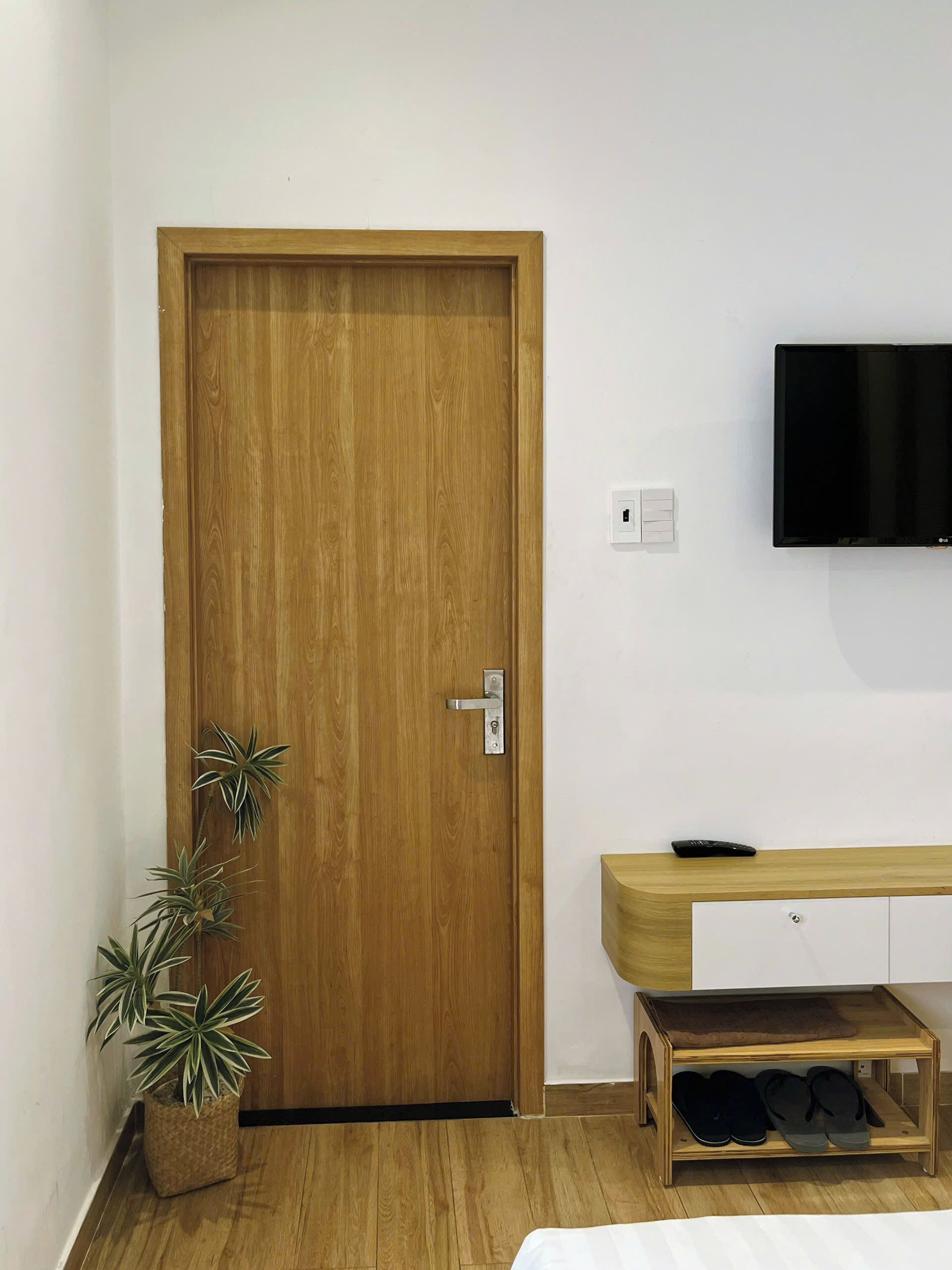 A room with a wooden door, a mounted TV, a wooden bench with a shoe rack, and a potted plant.