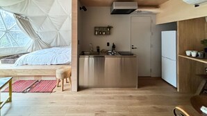 A compact kitchen with a central island, a built-in sink, and a refrigerator. There is a window with a view of trees, a bed with a white canopy, and a small stool. - villa Fukuza (Naganuma)
